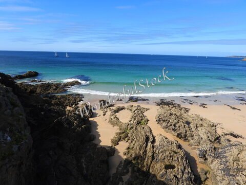 Plage de sable et rochers à Plouzané, Bretagne, France, avec des voiliers au loin et une mer turquoise sous un ciel dégagé.