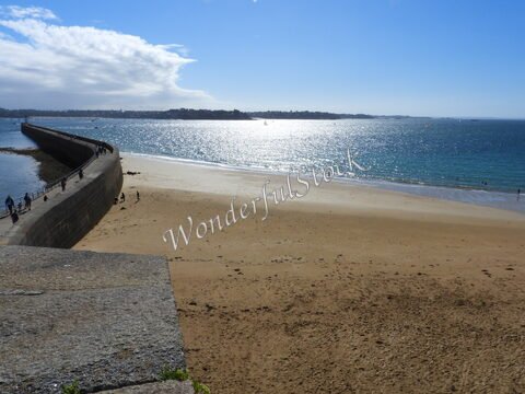 Plage de Saint-Malo en Bretagne, France, avec un mur en pierre et des personnes se promenant le long de l'eau scintillante.