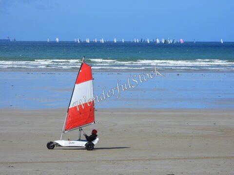 Véhicule à voile sur la plage de Sainte-Anne-la-Palud, en Bretagne, avec des voiliers en arrière-plan sur l'océan.