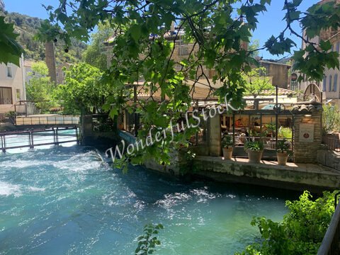 Fontaine et restaurant à Fontaine du Vaucluse, France, avec un cours d'eau et des arbres en arrière-plan.