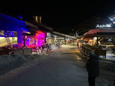 Rue animée et éclairée de La Plagne, France, avec des terrasses de restaurants et un décor hivernal.