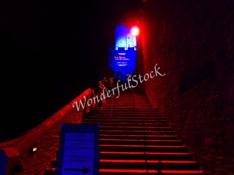 Escalier menant au Mont Saint-Michel, France, éclairé par des lumières rouges et bleues, avec des visiteurs montant les marches.