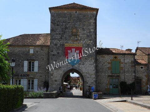 Arcade en pierre avec blason à Montpazier, France, entourée de maisons en pierre et de végétation.