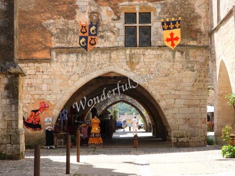 Des vêtements colorés exposés sous des arches en pierre à Montpazier, France, avec des blasons sur les murs.