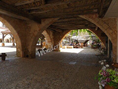 Place animée avec des arches en pierre à Montpazier, France, où des tables sont disposées sous des parasols.