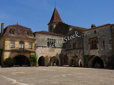 Village médiéval à Montpazier, France, avec des bâtiments en pierre et un clocher, sous un ciel bleu.