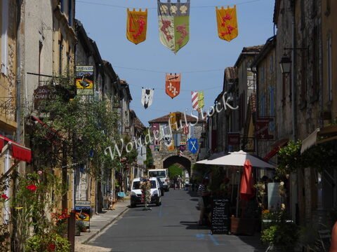 Rue animée de Montpazier, France, ornée de bannières colorées et de commerces, avec des maisons en pierre sur les côtés.