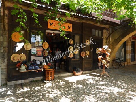 Vente de chapeaux devant un magasin à Montpazier, France, avec des chapeaux suspendus et un décor de pierres anciennes.