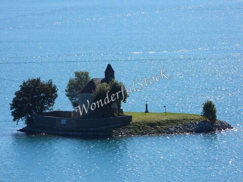 Église sur une île au lac de Serre-Ponçon, France, entourée d'eau turquoise et de quelques arbres.