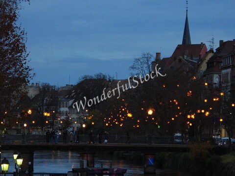 Pont illuminé au bord de l'eau à Strasbourg, France, avec des bâtiments en arrière-plan et des arbres décorés de lumières.