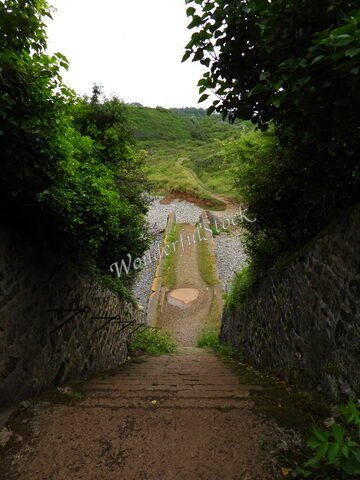 Un chemin de terre descendant entre des murs en pierre à Crozon, France, entouré de verdure et de galets.