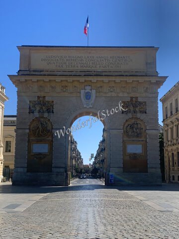 Arc de triomphe à Montpellier, France, avec un drapeau français flottant et des bâtiments historiques de part et d'autre de la rue pavée.