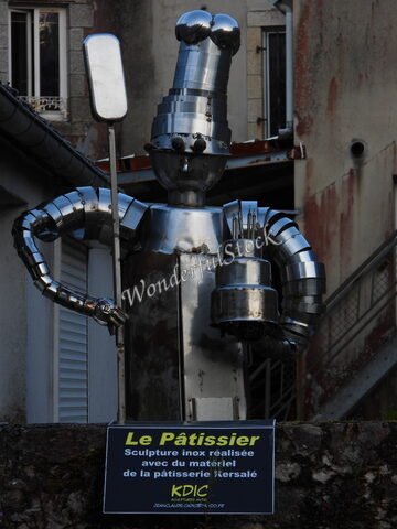 Sculpture d'un pâtissier en inox à Pont-Aven, France, avec des ustensiles de cuisine visibles et un fond d'immeubles.